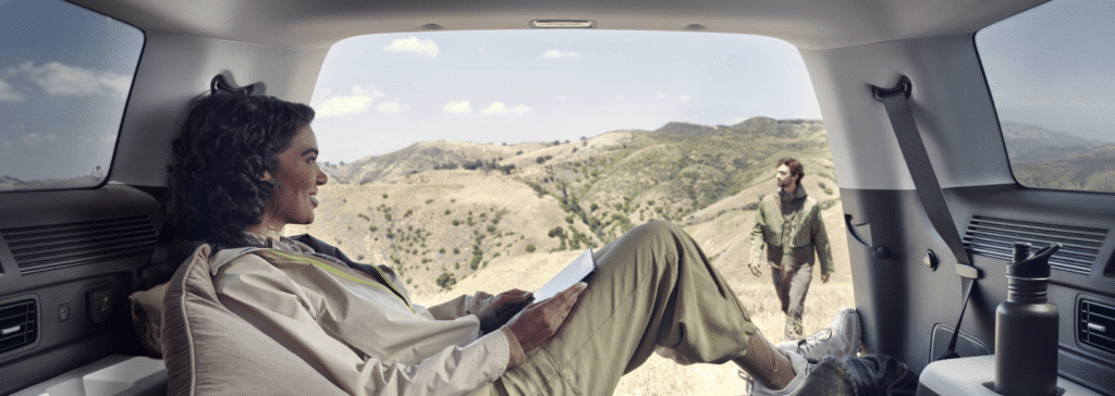 Person relaxing inside the back of a car, looking out at another person walking in a scenic, mountainous landscape.